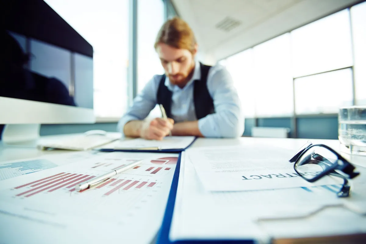 businessman sitting at a desk in a bright office, reviewing and signing financial documents.